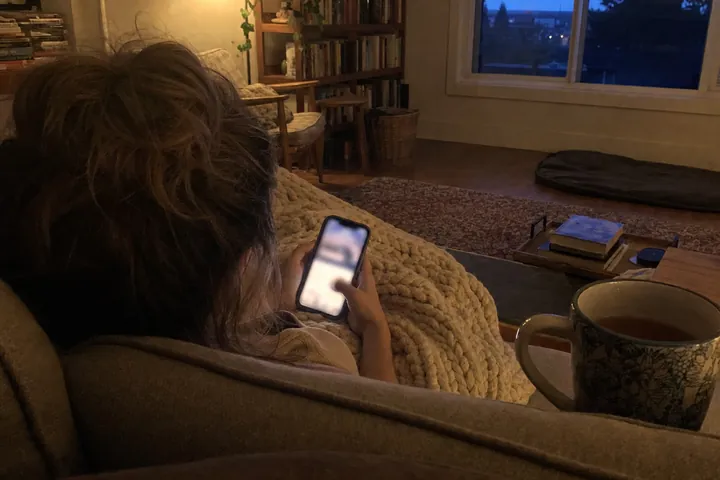 Seen from behind, a person with a messy bun sits on a sofa under a chunky knit blanket in a dim living room at dusk, looking at a phone, with a mug of tea and bookshelves nearby.