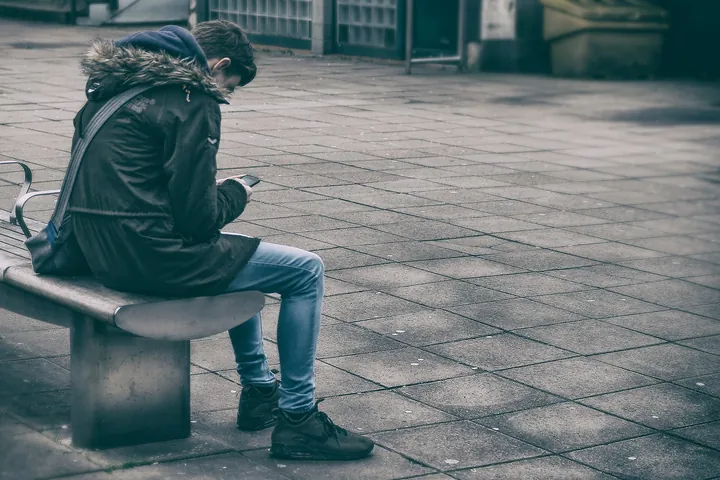 A person in a dark hooded jacket and jeans sits alone on a bench on an empty paved square, head down over a phone; the face is hidden by the hood.