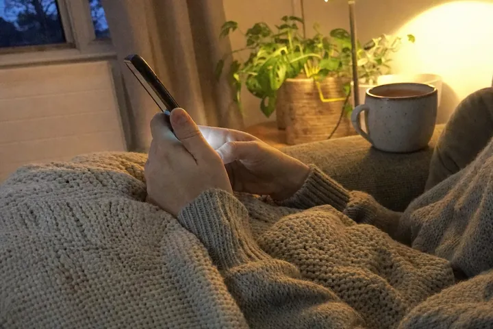 Two hands hold a phone on a lap under a knitted throw on a sofa in the evening, a speckled mug of tea on the armrest and a lamp glowing behind.
