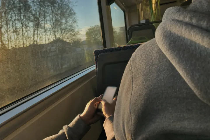 Seen from behind, a person in a grey hoodie on a commuter train types on a phone with both thumbs, low golden sun coming through the window.