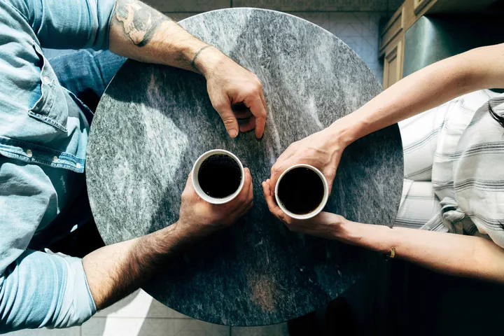 Seen from above, two people sit across a round marble café table, each holding a black coffee; only their hands and arms are visible.