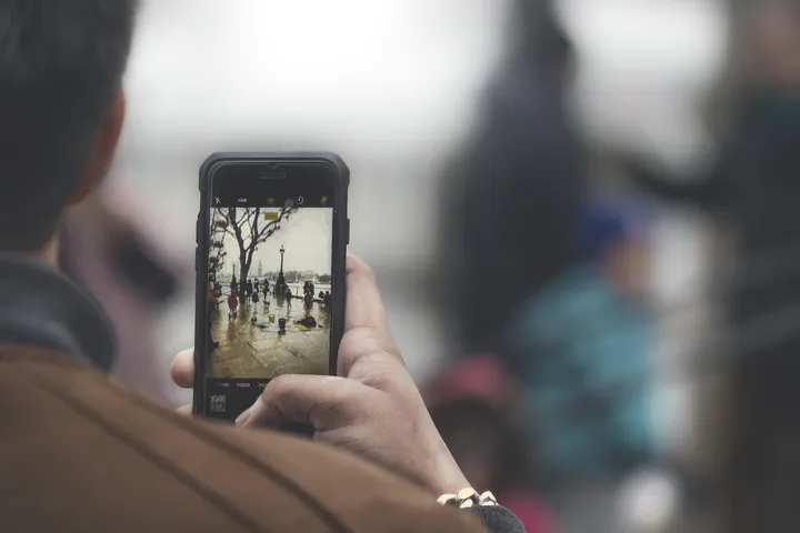 Over-the-shoulder view of a person in a brown jacket holding up a smartphone to photograph a wet riverside street with bare trees and people; the person faces away.