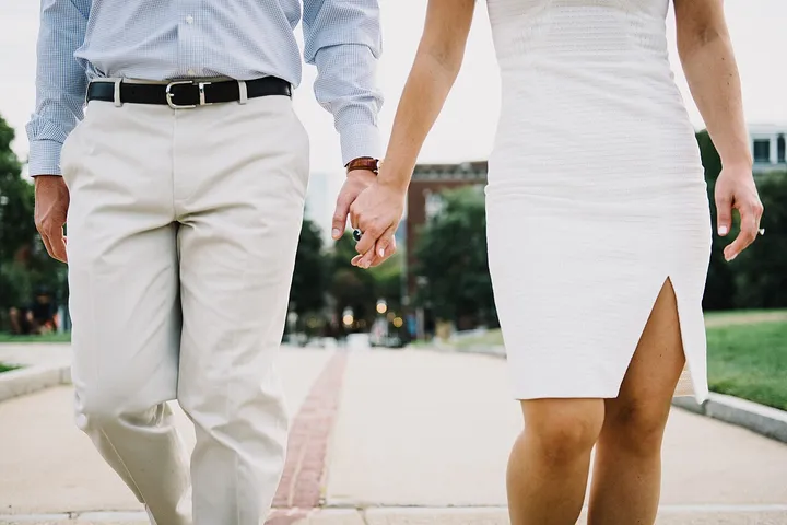 Two people holding hands while walking along a city path, cropped at the shoulders so only their hands, arms and clothes are visible.