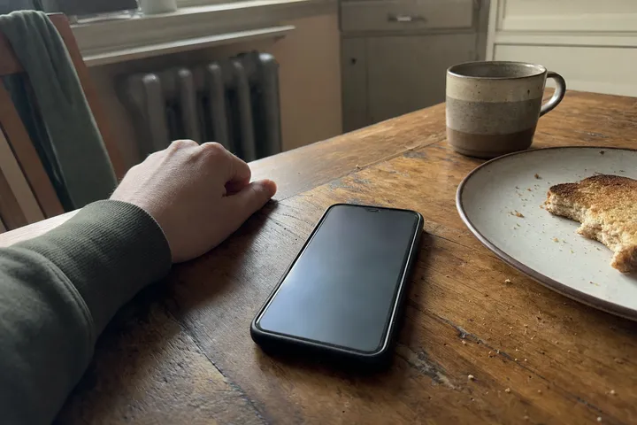 A hand resting on a worn wooden kitchen table next to a phone with a dark screen, a ceramic mug and a plate with half-eaten toast in morning light.
