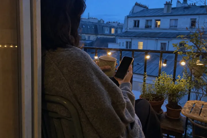 Seen from behind, a person in a grey cardigan sits on a small city balcony at blue hour holding a phone and a mug, with string lights and blurred rooftops beyond.
