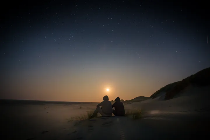 Two people in hooded tops sit side by side on a sand dune at night, seen from behind, watching a low moon under a starry sky.
