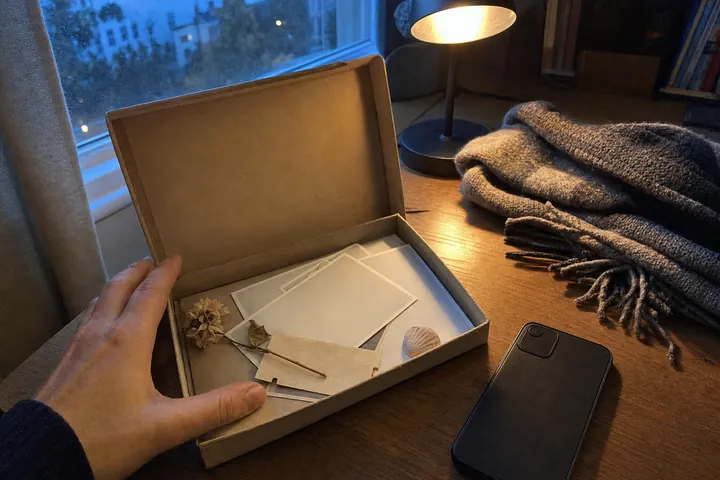 An open cardboard keepsake box on a wooden desk holding blank photo prints, a dried flower, a ticket and a seashell, with a phone lying screen-down beside it under a warm lamp at dusk.