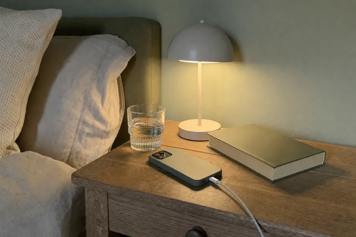 A phone charging face down on a wooden bedside table under a warm lamp, next to a glass of water and a folded book.
