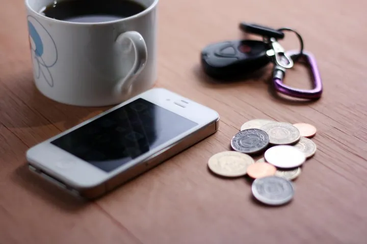 An older white smartphone lying on a wooden table next to a small pile of coins, a coffee mug and a set of keys.