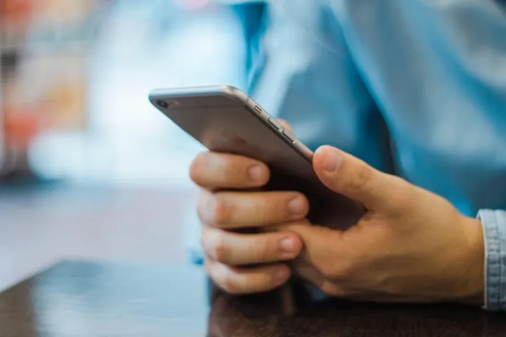 A pair of hands holding an iPhone over a wooden table, with the screen facing away from the camera.